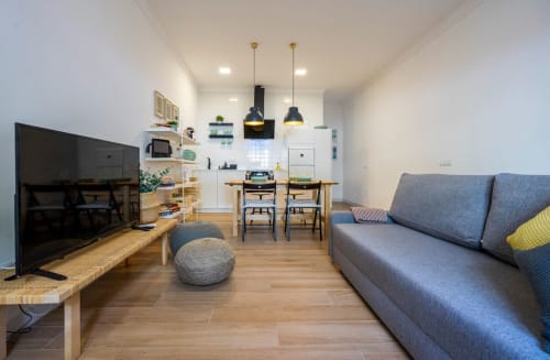 Área de estar de apartamento moderno de planta abierta en Nerja con sofá azul, TV, mesa de comedor y cocina con luces colgantes