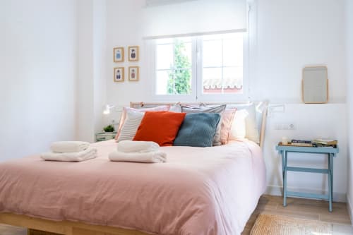 Dormitorio luminoso en apartamento de Nerja con ropa de cama rosa, cojines coloridos y mesita azul junto a ventana con vistas a plantas