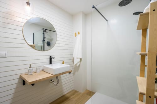 Cuarto de baño moderno en Apartamentos Minaga de Nerja con paredes de tablillas blancas, espejo redondo, tocador de madera y ducha tipo lluvia