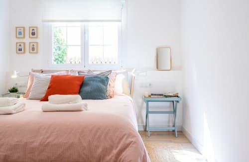 Dormitorio luminoso en apartamento de Nerja con ropa de cama rosa, almohadas coloridas, vista por ventana y mesita azul