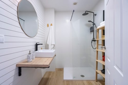Baño moderno en apartamento de Nerja con azulejos blancos, espejo redondo, tocador de madera y ducha a ras de suelo con accesorios negros
