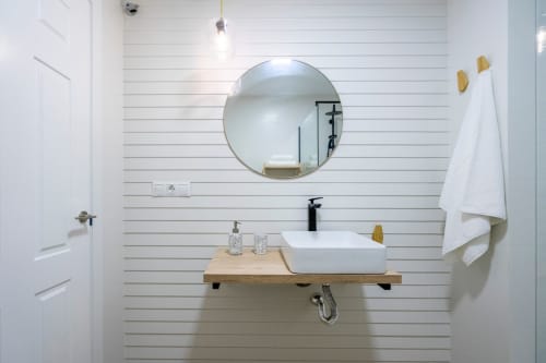 Baño moderno en apartamento de Nerja con espejo redondo, lavamanos blanco, estante de madera y paredes de tablas