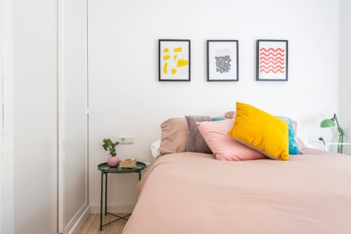 Dormitorio luminoso en apartamento de Nerja con ropa de cama rosa, almohada amarilla y tres cuadros en pared blanca sobre la cama