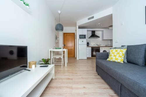 Moderno salón en Nerja con sofá gris, cocina americana, televisión y suelos de madera