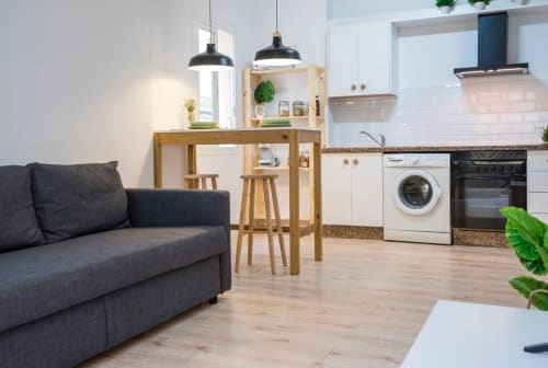 Apartamento moderno de planta abierta en Nerja con sofá oscuro, mesa de comedor de madera, cocina blanca con electrodomésticos y lámpara colgante negra