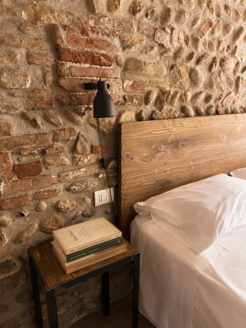 Schlafzimmer in den Apartments Borgovivobg mit freiliegenden Backsteinmauern, Holzbettrahmen, Nachttisch mit Büchern und Leseleuchte