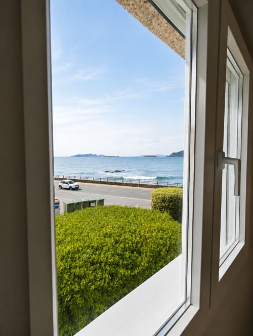 Vista abierta del frente marítimo de Baiona con jardín verde, coche estacionado y aguas de la bahía azul desde el balcón del apartamento As Estelas