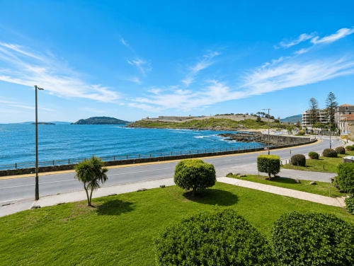 Paseo costero de Baiona con jardines cuidados, vistas al mar azul y fortaleza histórica en la ladera