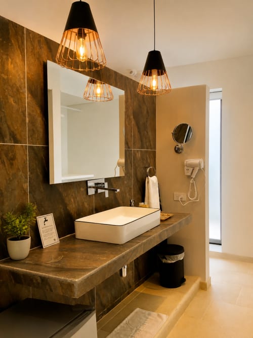 Baño moderno en La Masia Hotel Boutique con paredes de mármol oscuro, lavamanos blanco, espejo iluminado y lámpara colgante dorada