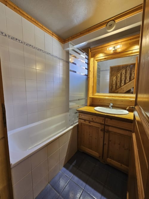 Salle de bain aux murs carrelés blancs, meuble-lavabo en bois, lavabo rond, miroir, baignoire avec paroi vitrée, sol sombre aux Étoiles de