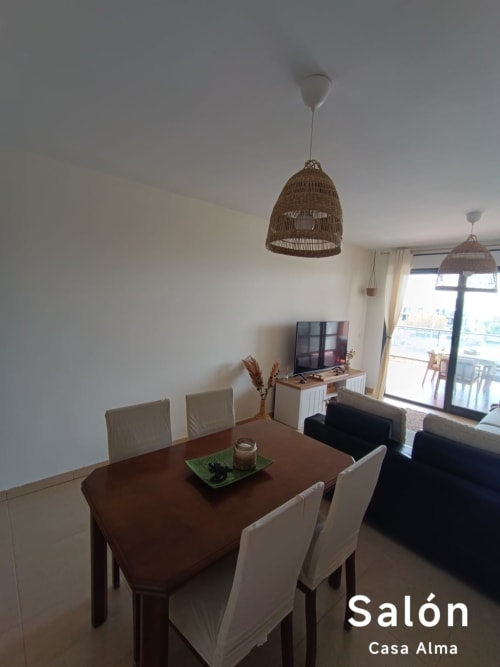 Salle à manger moderne de l'appartement Casa Alma avec table en bois, chaises beige, luminaires suspendus tissés et vue sur le balcon à Sant