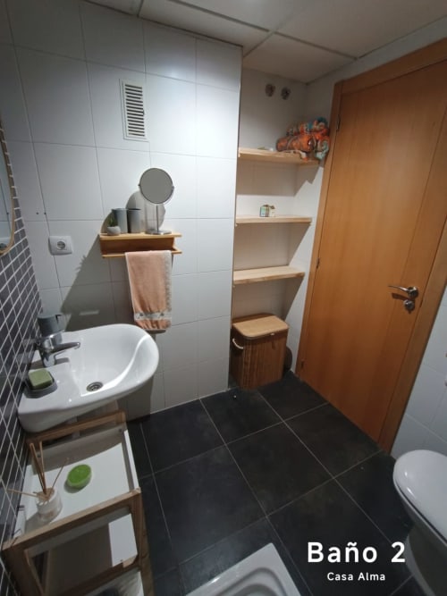 Salle de bains moderne avec carrelage blanc, sol noir, lavabo, toilettes, étagères en bois, miroir et porte-serviettes à l'appartement Casa Alma
