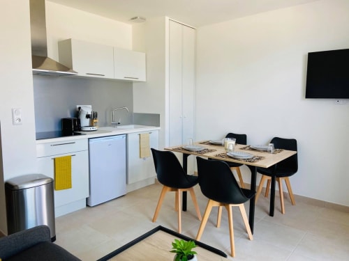 Appartement T2 moderne à la Résidence Marina di Bravone avec cuisine ouverte, coin repas aux chaises noires et TV murale