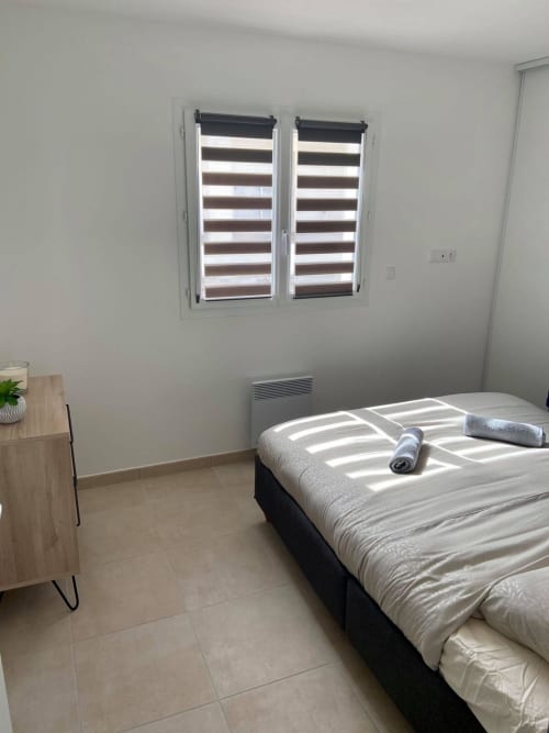 Chambre moderne à Marina di Bravone avec stores rayés, lit avec serviette roulée, table de nuit en bois et sol carrelé