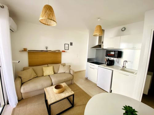 Modern studio apartment at Résidence Marina di Bravone with beige sofa, kitchenette, woven pendant lights, and minimalist