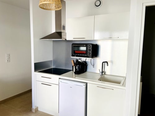 Modern white kitchenette with black cooktop, oven, sink, and woven pendant light at Résidence Marina di Bravone, Linguizzetta
