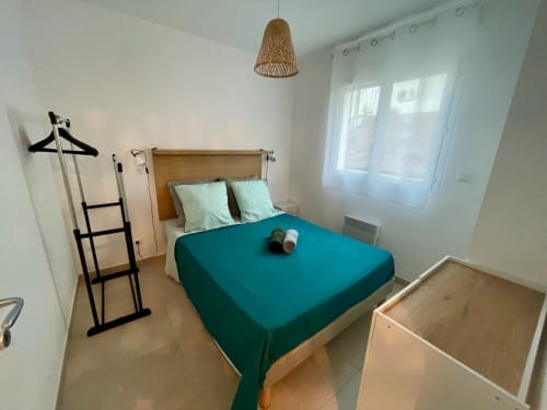 Chambre lumineuse à la Résidence Marina di Bravone avec literie bleu-vert, tête de lit en bois, lampe suspendue en rotin et portes-fenêtres