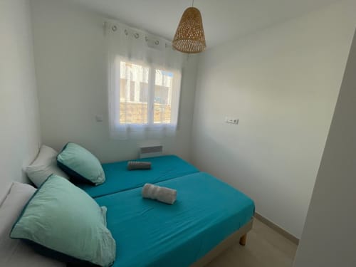 Camera da letto luminosa a Marina di Bravone con letto turchese, cuscini menta, lampada sospesa intrecciata e finestra con vista su Linguizzetta