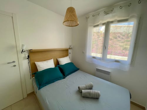 Chambre moderne à la Résidence Marina di Bravone avec tête de lit en bois, oreillers bleu-vert, luminaire suspendu tressé et vue sur mer