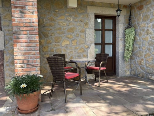 Rustic patio at El Picoretu with stone walls, wicker chairs with red cushions, potted flowers, and mountain views near Mestas