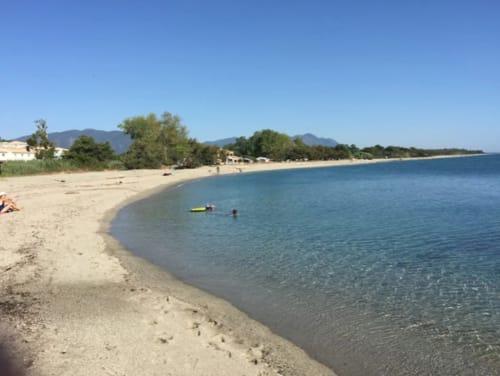 Sandy beach at Marina di Bravone in Corsica with turquoise water, swimmers, mountains in distance, and beachfront buildings