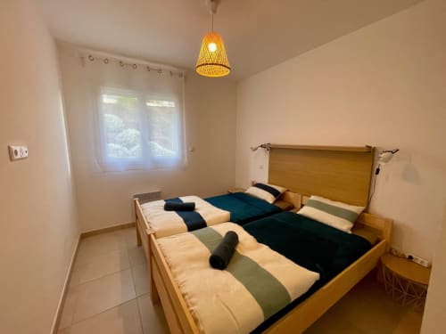 Chambre lumineuse avec deux lits en bois à la Résidence Marina di Bravone, Linguizzetta. Murs crème, lampe suspendue, fenêtre avec