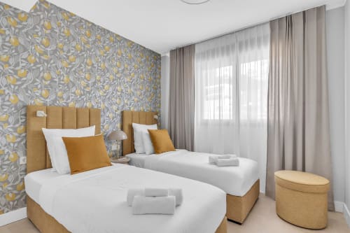 Nowoczesna sypialnia z dwoma łóżkami w apartamencie HomyMarbella w Elvirii ze złotymi zagłówkami, żółtą tapetą w deseń i widokiem na ogród