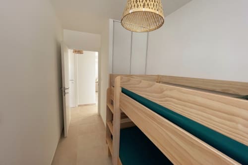 Chambre lumineuse à la Résidence Marina di Bravone avec lit à cadre bois, matelas bleu-vert, lampe au plafond tissée et porte ouverte.