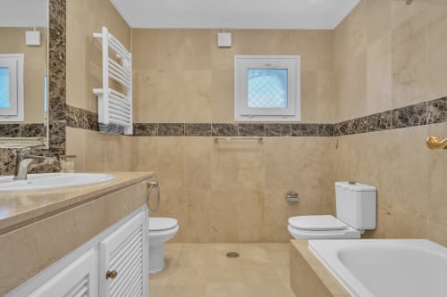Elegant bathroom at Casa Perla Marbella with beige marble tiles, white fixtures, bathtub, and decorated border trim