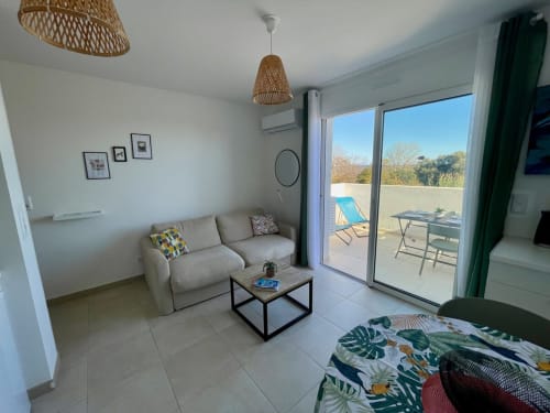 Moderno appartamento presso Résidence Marina di Bravone con divano beige, lampade a sospensione in vimini e accesso alla terrazza con vista sul mare