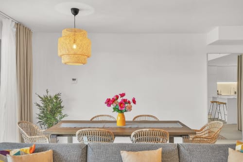 Nowoczesna jadalnia w townhouse HomyMarbella z plecioną lampą wiszącą, drewnianym stołem, rattanowymi krzesłami i różowymi kwiatami