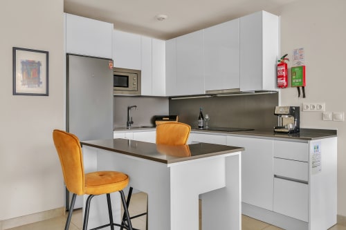 Nowoczesna kuchnia z białymi szafkami, ciemnymi blatami i pomarańczowymi krzesłami przy wyspie w apartamencie HomyMarbella, Elviria