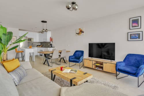 Nowoczesny salon apartamentu w Marbelli z niebieskimi aksamitnymi fotelem, drewnianym stojakiem TV, jadalnią, kuchnią i widokami na góry