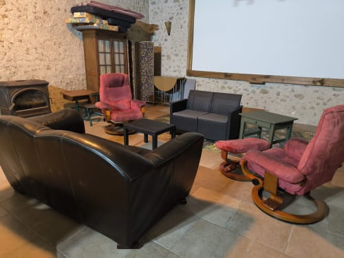 Salon rustique du Gîte de la vallée avec canapé en cuir noir, fauteuils rose, cheminée et vue sur le jardin