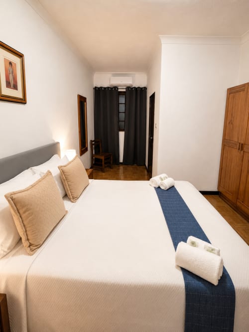 Moderno quarto duplo na Serrana Gerês Inn com roupa de cama branca, almofadas bege, caminho de cama azul e detalhes em madeira