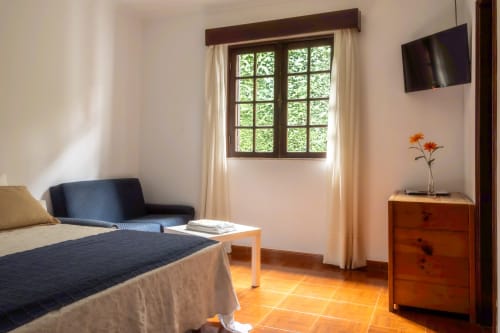 Quarto Duplo Luminoso na Pousada Serrana Gerês com cama, poltrona, cómoda de madeira e vistas para o jardim pela janela ampla