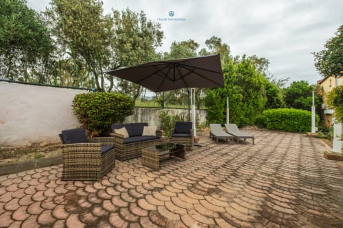 Patio esterno di House Live Express con sedute in vimini, ombrellone nero e lettini che si affacciano sui giardini di Quartu