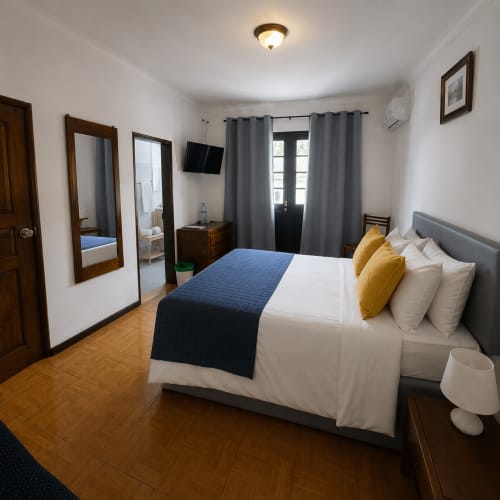 Quarto duplo da Pousada Serrana Gerês com cama estofada cinzenta, roupa de cama branca, almofadas amarelas, piso em madeira e varanda com