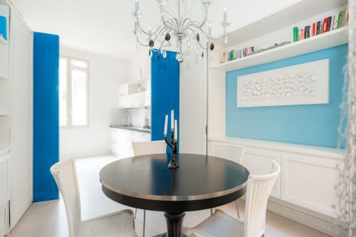 Zona pranzo moderna con tavolo rotondo nero, sedie in vimini bianco, parete azzurra, lampadario e cucina a Perla Marina