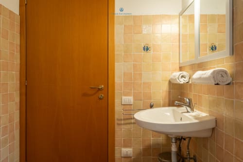 Bagno con piastrelle beige, lavabo bianco, specchio e asciugamani arrotolati presso l'hotel House Live Express a Quartu Sant'Elena