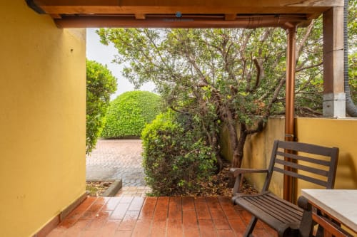 Patio coperto di House Live Express con sedia in legno, pavimento in cotto rosso e vista sul lussureggiante giardino a Quartu Sant'elena
