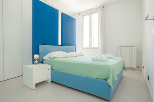 Camera da letto moderna e luminosa con parete blu, biancheria verde, comodino bianco e vista sulla città a Perla Marina, Finale Ligure