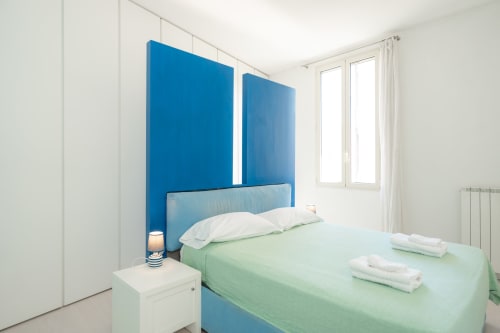 Camera da letto moderna con testata azzurra, biancheria verde, cuscini bianchi, lampada comodino e vista sulla città a Perla