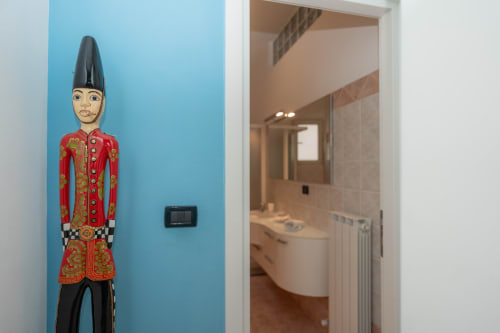 Figura decorativa in legno con abito rosso su parete blu, bagno visibile dalla porta dell'appartamento Perla Marina