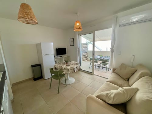 Luminoso soggiorno a Résidence Marina di Bravone con divano beige, tavolo da pranzo, angolo cottura e accesso al balcone