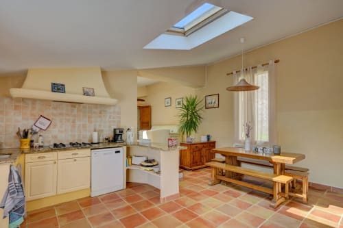 Cuisine-salle à manger lumineuse du gîte Lavande avec placards crème, carrelage en terre cuite, table en bois, puits de lumière et jardin