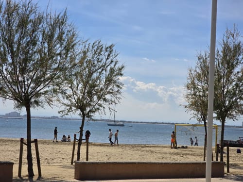 Sandy beach in Rosas with trees, volleyball net, and sailboat on blue water near family apartment