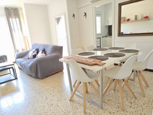 Modern dining area with white chairs and wooden table in family apartment near Rosas beach, with blue futon and bright living