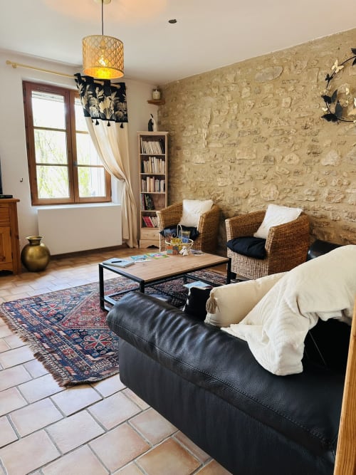 Spacieux séjour du Gîte Romarin avec canapé en cuir noir, fauteuils en osier, murs en pierre et carrelage en terre cuite