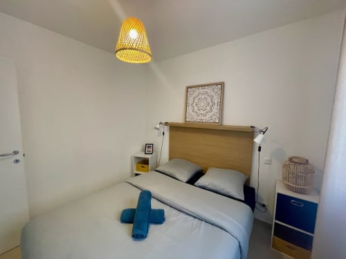 Modern bedroom at Résidence Marina di Bravone with wooden bed, yellow pendant lamp, blue nightstand, and white walls in